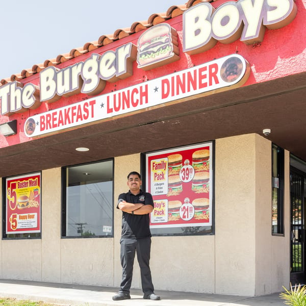 A man in a black polo shirt stands proudly in front of The Burger Boys restaurant.