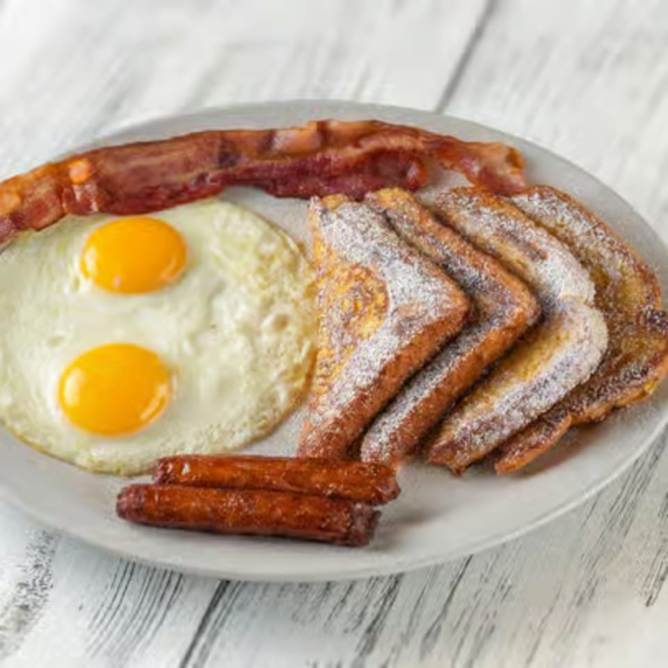 A breakfast plate with two fried eggs, bacon, sausage links, and powdered sugar French toast.