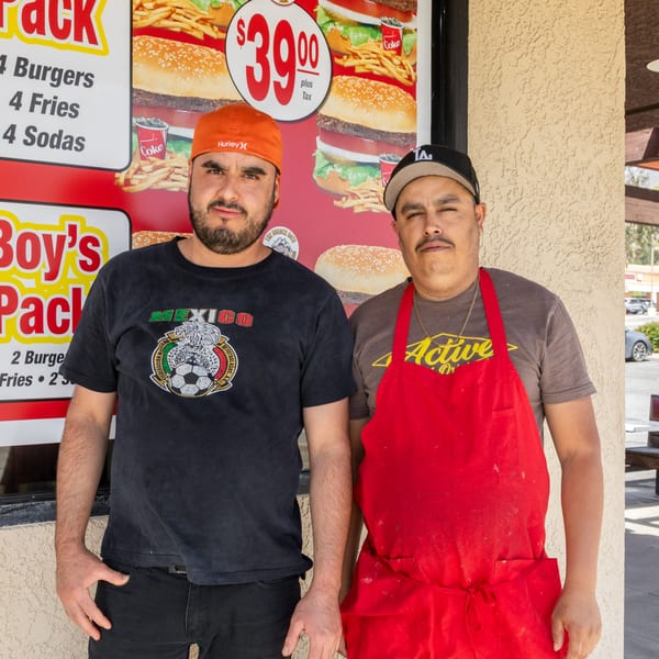 Two men stand in front of a fast-food restaurant with a menu featuring burgers, fries, and sodas.